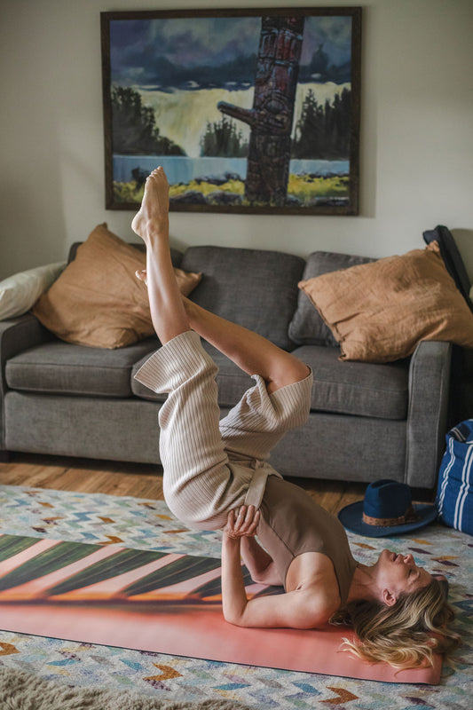 Person doing a yoga pose on a mat in a living room with a couch and painting in the background.
