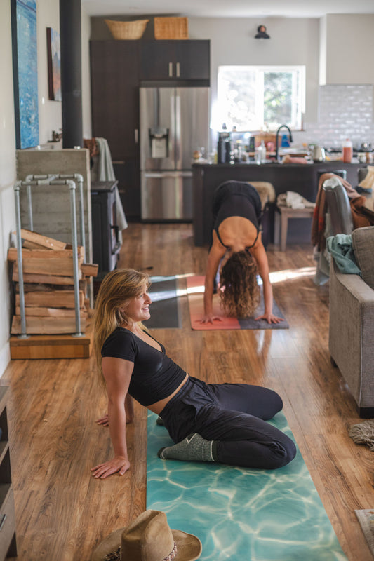 Two women exercising on yoga mats in a home setting with a pool effect on the floor.