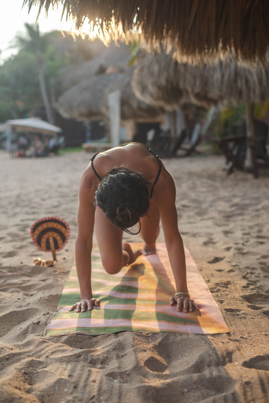 Person practicing yoga on a colorful mat on a sandy beach with thatched structures in the background.