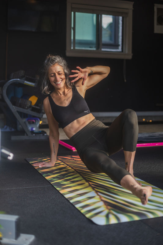 Woman in black sports bra and green leggings on a colorful yoga mat in a home setting.