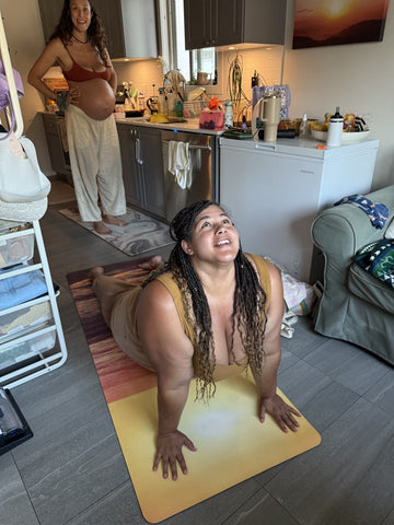 Two women in a kitchen, one on a yoga mat, with a sunset painting on the wall.